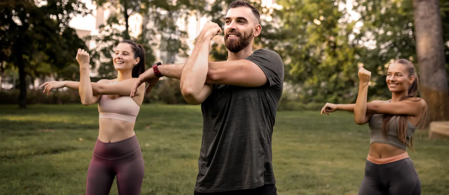 Gruppe von drei Personen in Sportkleidung dehnt Arm- und Schultermuskeln auf einer Grünfläche im Freien. Im Hintergrund stehen hohe Bäume.