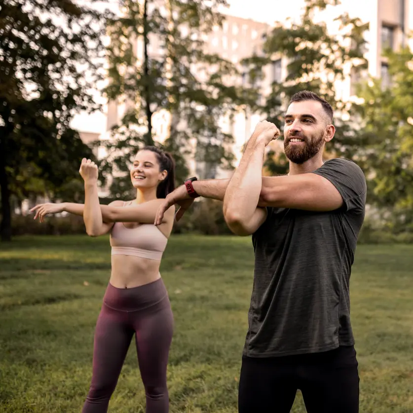 Gruppe von drei Personen in Sportkleidung dehnt Arm- und Schultermuskeln auf einer Grünfläche im Freien. Im Hintergrund stehen hohe Bäume.