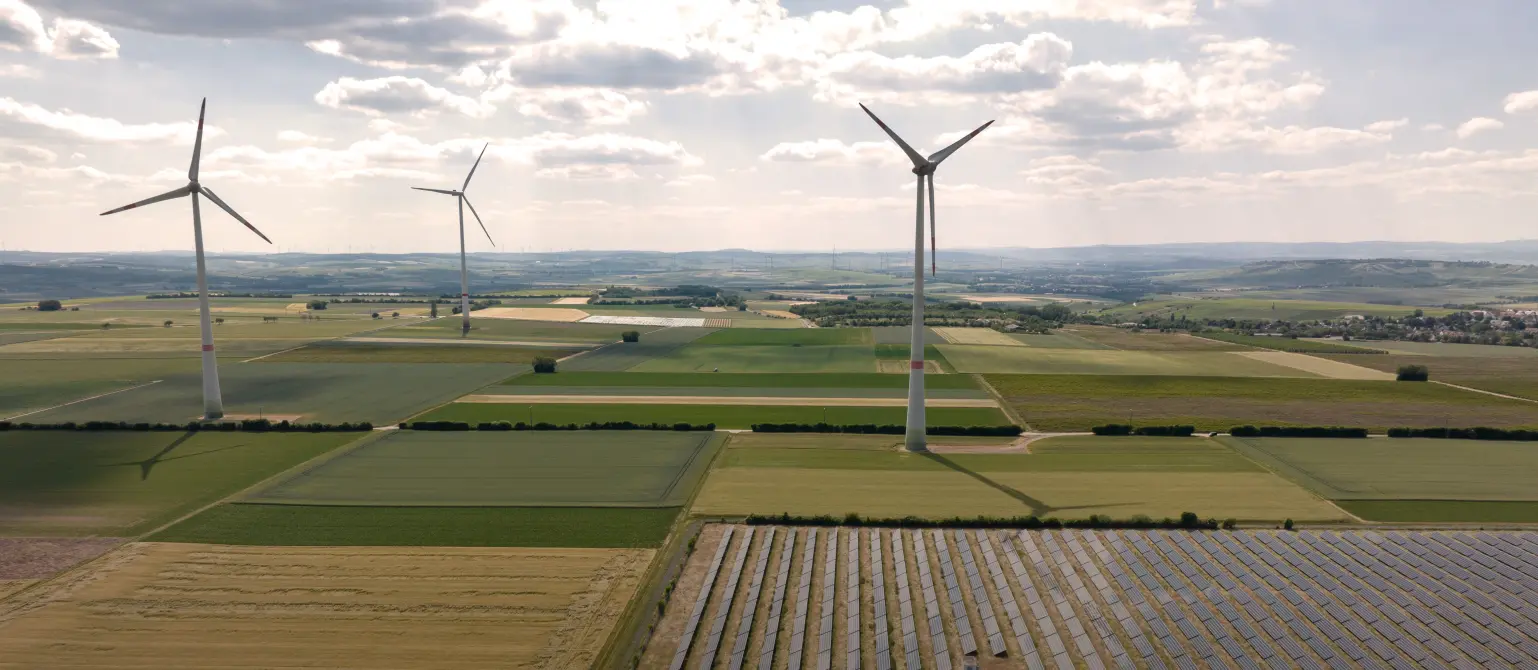 Luftaufnahme eines Solarparks und mehrerer Windkraftanlagen im Hintergrund in einer weiten flachen Agrarlandschaft.