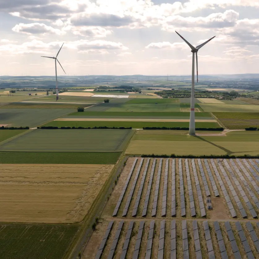 Luftaufnahme eines Solarparks und mehrerer Windkraftanlagen im Hintergrund in einer weiten flachen Agrarlandschaft.