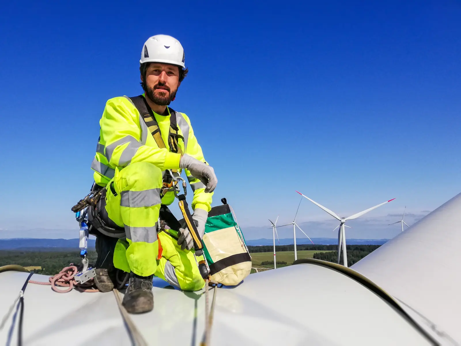 Fachkraft in Sicherheitsausrüstung arbeitet auf einer Windkraftanlage mit weiteren Windrädern im Hintergrund unter blauem Himmel.