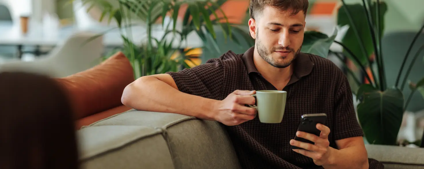 Junger Mann sitzt auf dem Sofa in einem modernen Lounge‑Bereich, umgeben von Pflanzen. Er hält eine grüne Tasse und schaut auf sein Smartphone. 