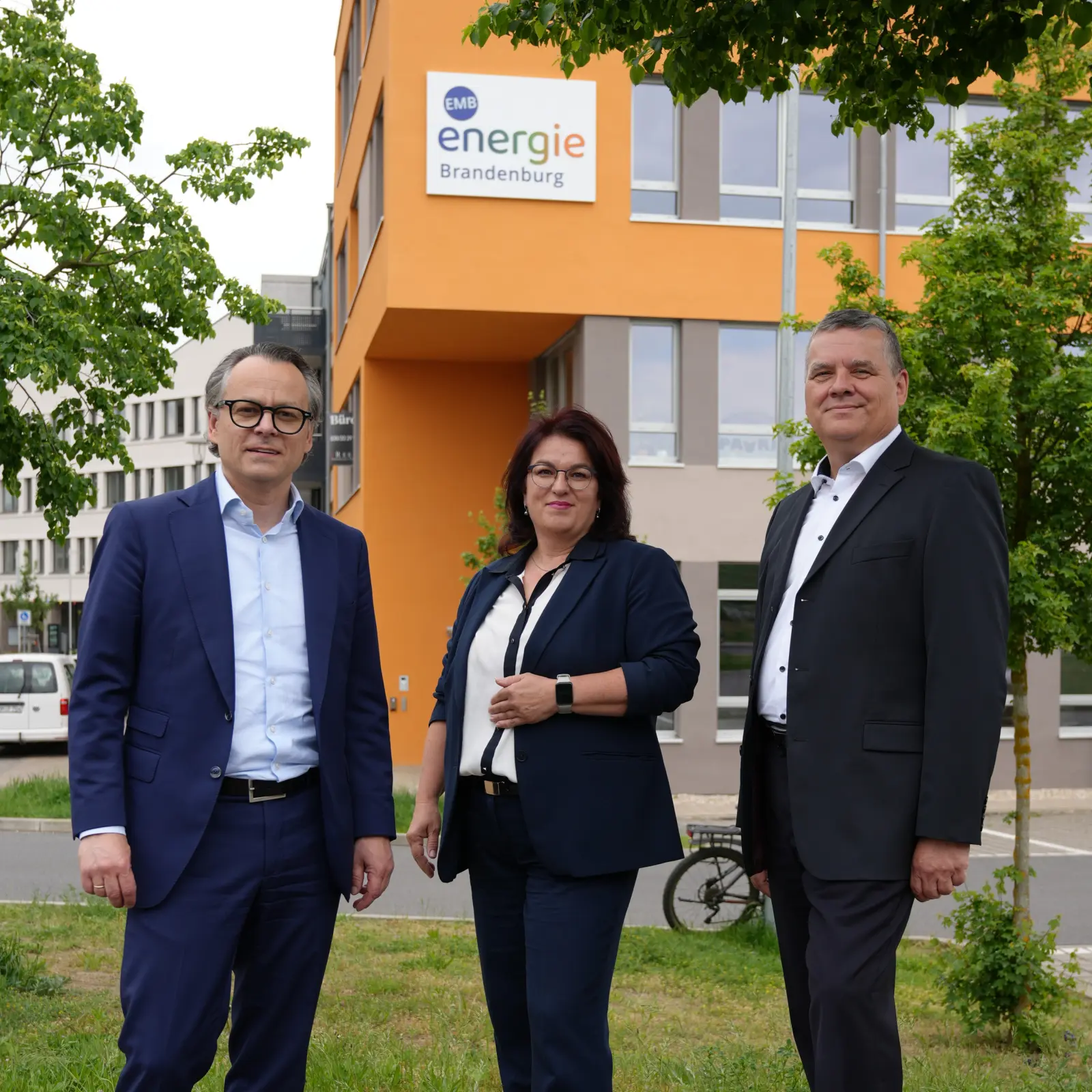 Drei Personen in Businesskleidung stehen vor einem Gebäude mit Energie Brandenburg-Logo im Außenbereich.