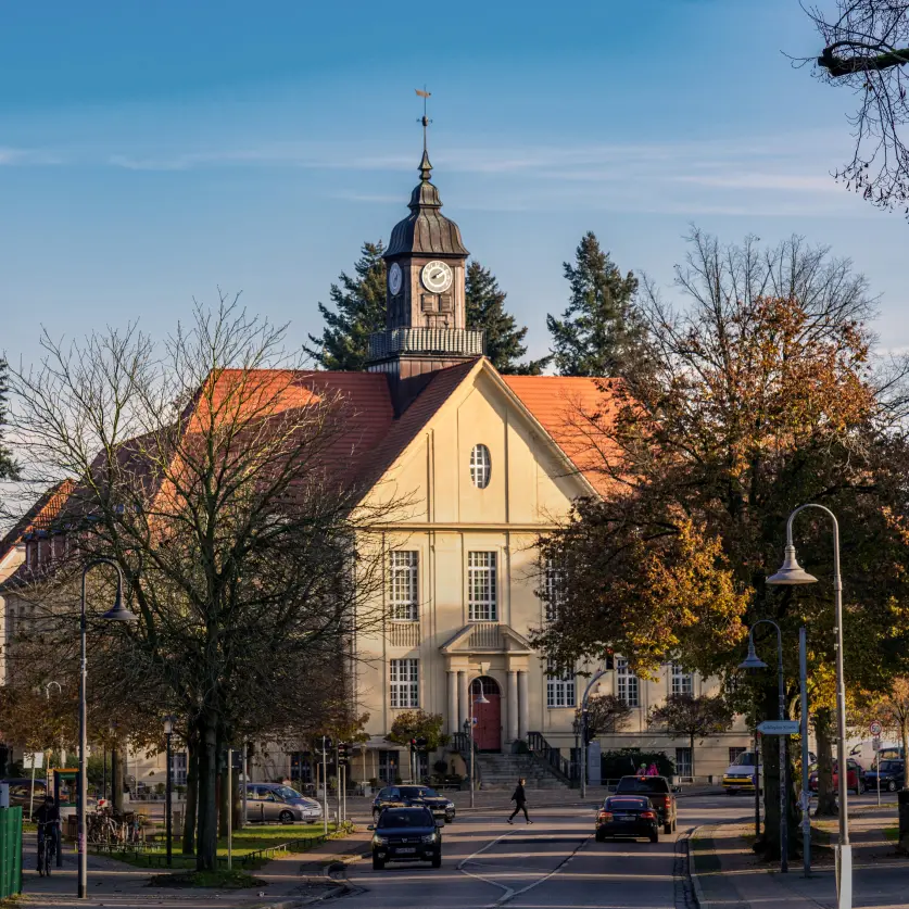 Historisches Rathaus von Birkenwerder mit Uhrturm an einer belebten Straße, umgeben von Bäumen und Wohnhäusern.