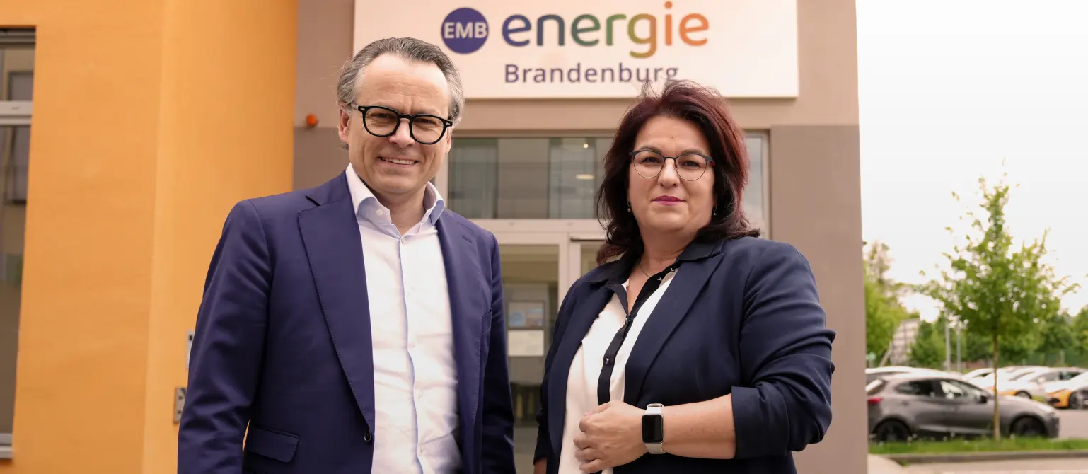 Leif Christian Cropp und Kathrin Jung: Zwei Personen in Businesskleidung stehen vor dem Eingangsbereich eines Gebäudes mit Energie Brandenburg-Logo.