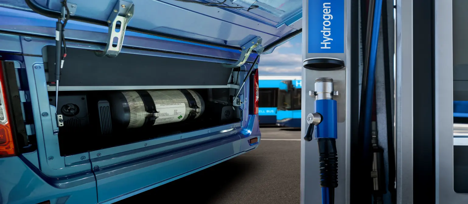 Ein blauer Bus mit geöffneter Heckklappe, in der man einen Wasserstofftank sieht, steht an einer Wasserstoffzapfsäule an einer Wasserstofftankstelle.