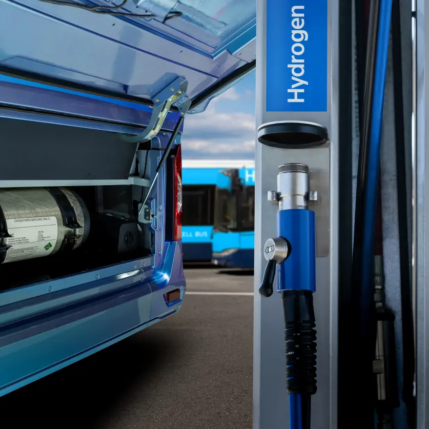 Ein blauer Bus mit geöffneter Heckklappe, in der man einen Wasserstofftank sieht, steht an einer Wasserstoffzapfsäule an einer Wasserstofftankstelle.