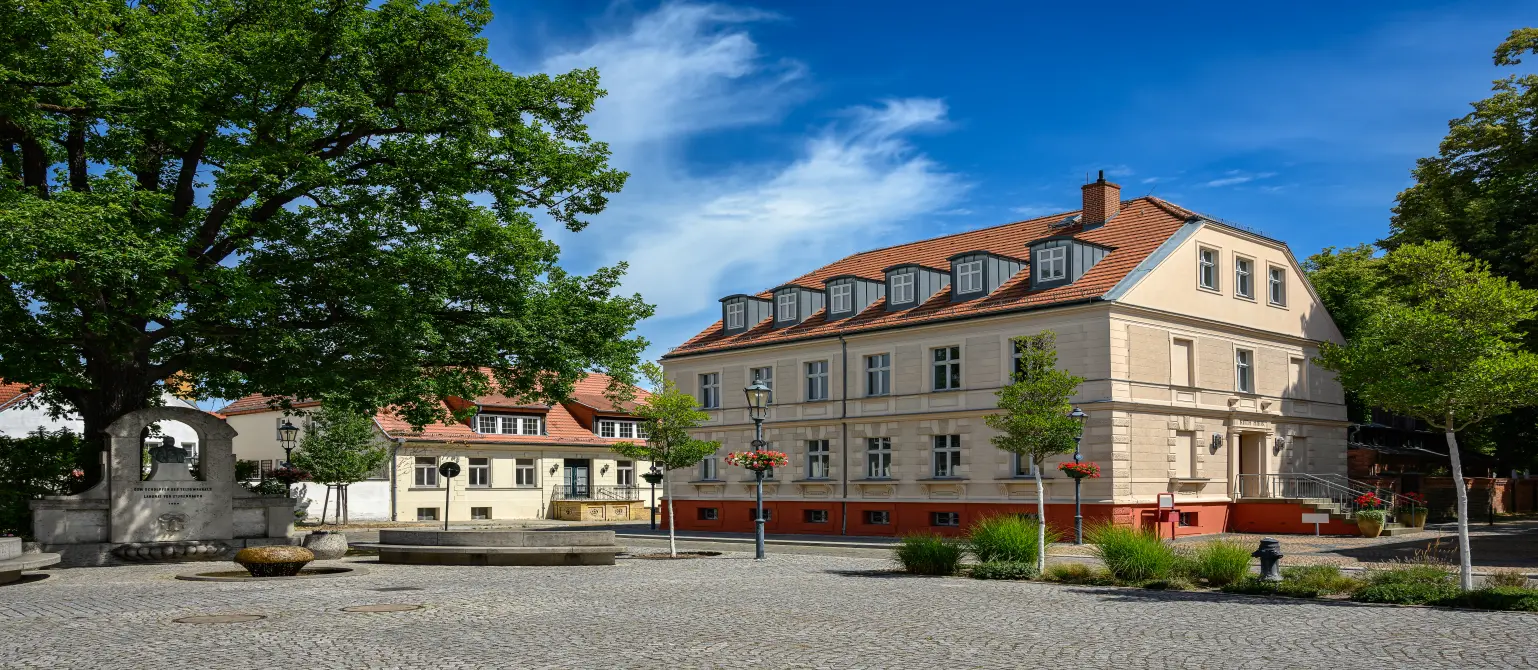 Das alte Rathaus der Stadt Teltow im Land Brandenburg mit rotem Walmdach. Auf dem gepflasterten Vorplatz plätschert ein Brunnen.