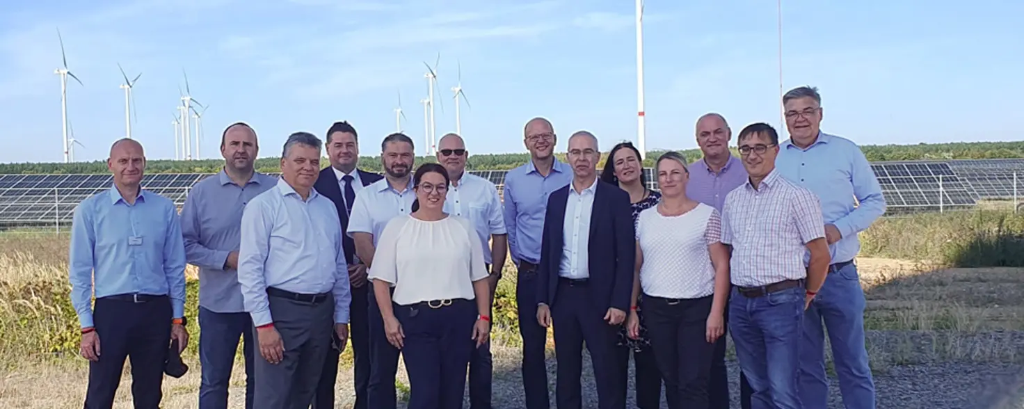 Eine Gruppe von Männern und Frauen in Business-Outfits stehen vor einem Photovoltaik-Park und Windkraftanlagen.
