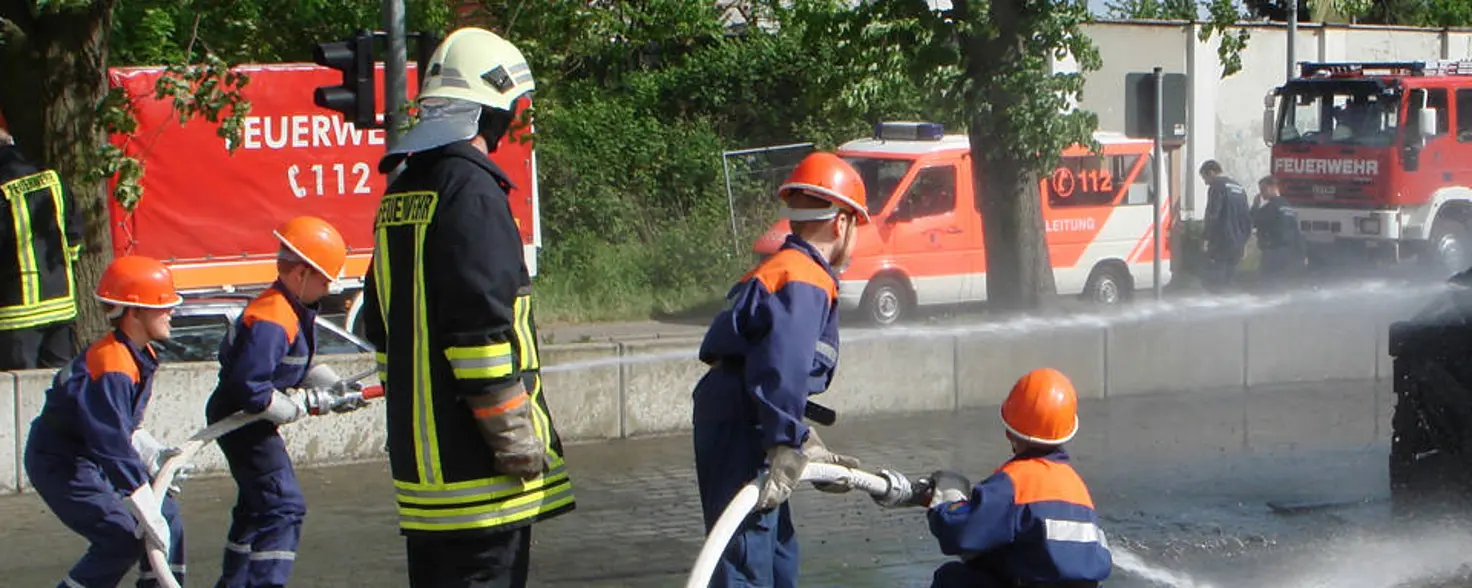 Vier Kinder in Schutzkleidung nehmen an einer Übung der Freiwilligen Feuerwehr Brandenburg teil und verspritzen Wasser mit Schläuchen.