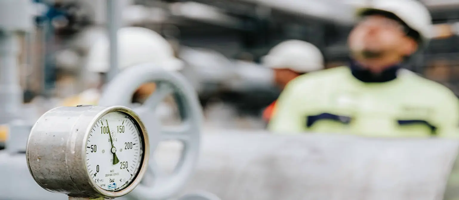 Eine Liquefied-Natural-Gas-Anlage (LNG). Im Vordergrund eine Druckanzeige, im Hintergrund sehr verschwommen Arbeiter mit Schutzhelm.