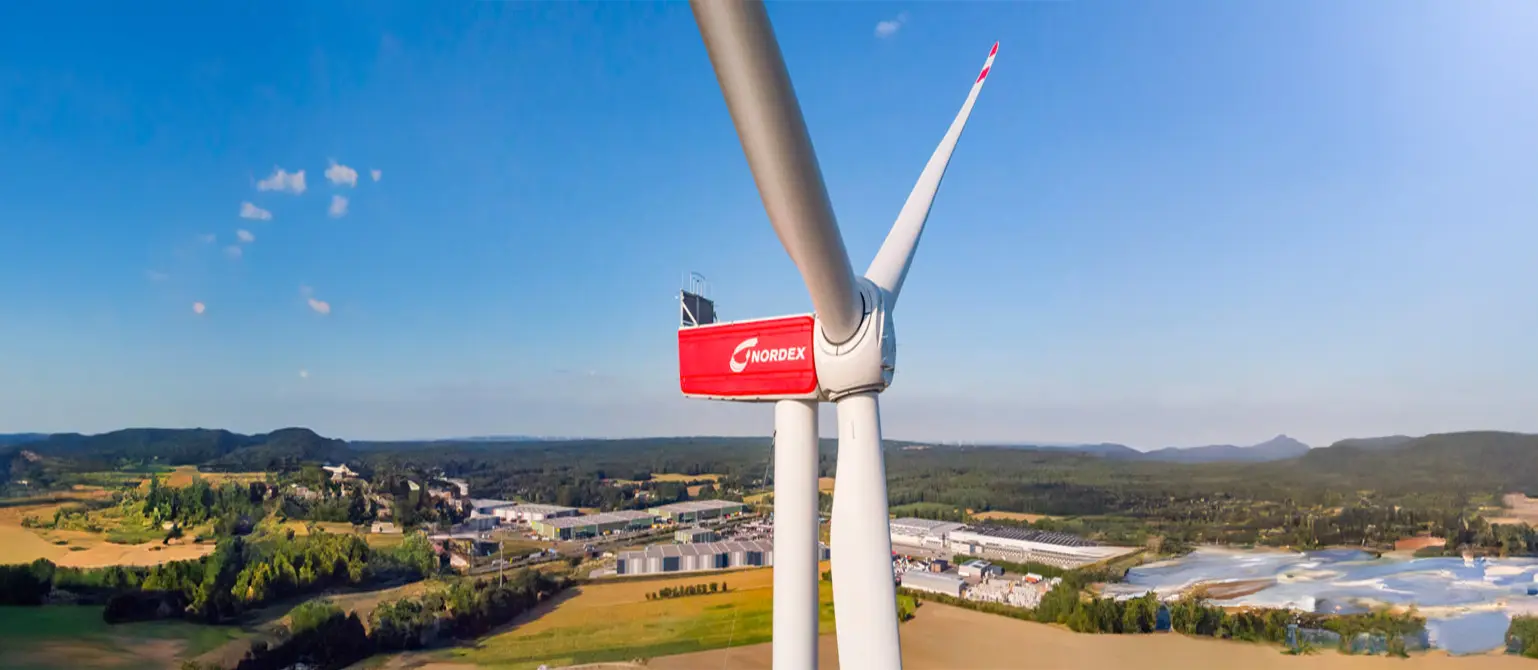 Eine rot-weiße Windkraftanlage von Nordex, aus der Höhe fotografiert und mit Fokus auf den Rotor. Im Hintergrund Wald und eine Stadt..