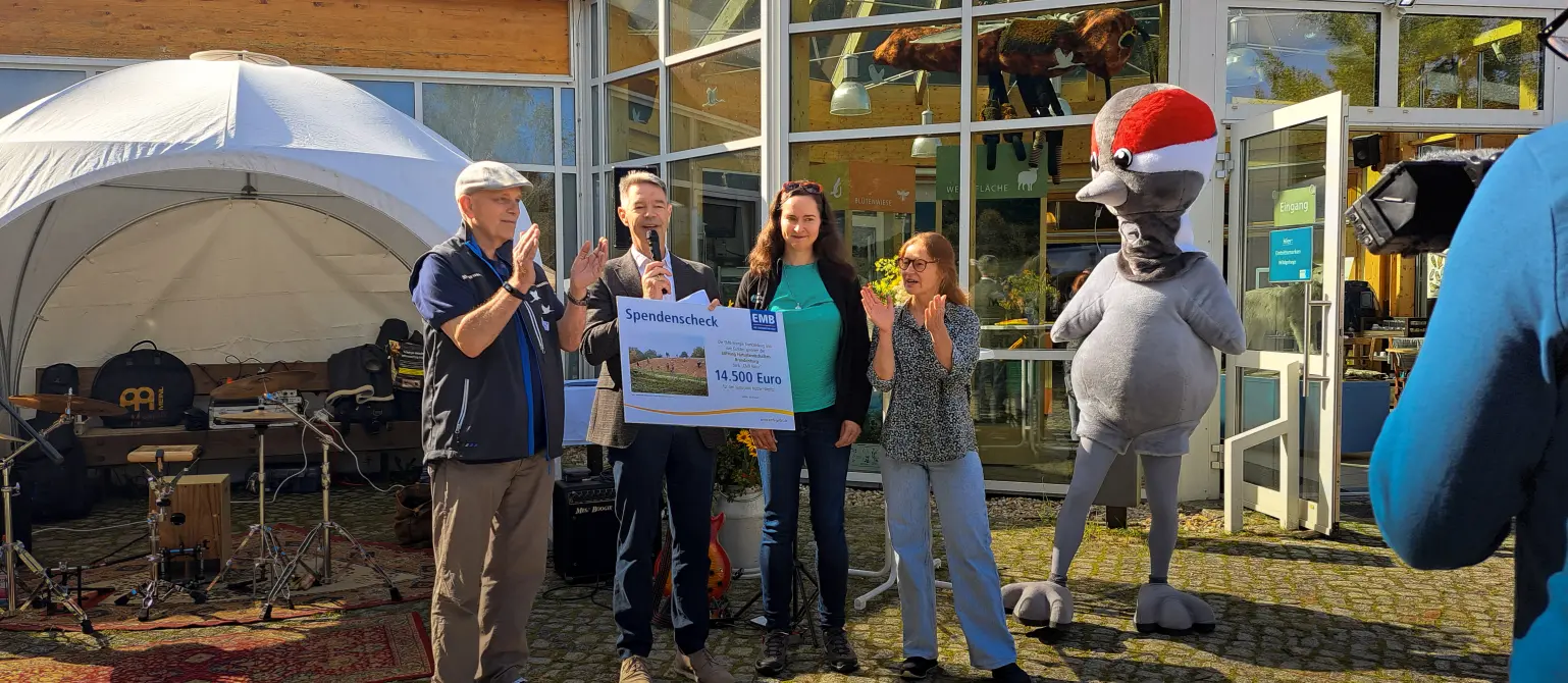 Ein Maskottchen und mehrere Personen halten einen Spendenscheck über 16.000 Euro. Im Hintergrund grüne Landschaft.