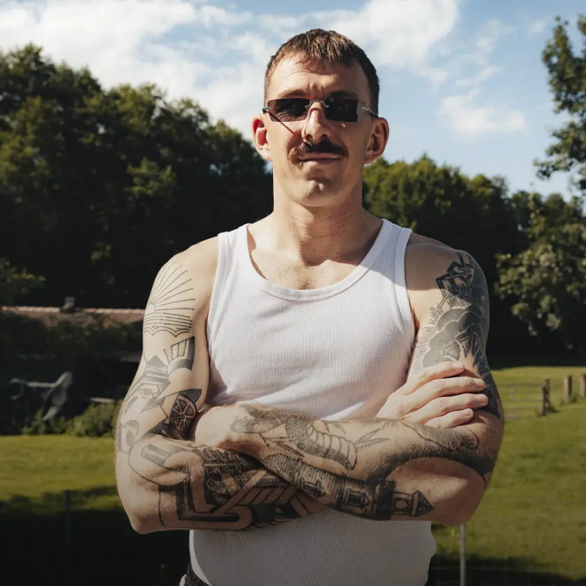 ättowierter Mann mit Schnurrbart Sonnebrille steht mit verschränkten Armen auf einer Wiese.