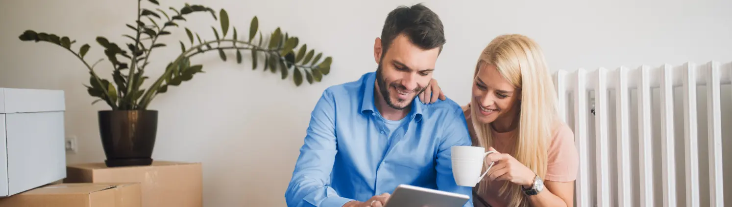 Glückliches Paar mit Tablet-Computer beim Kaffeetrinken auf dem Parkettboden seiner neuen Wohnung. Im Hintergrund Umzugskisten und ein Heizkörper.