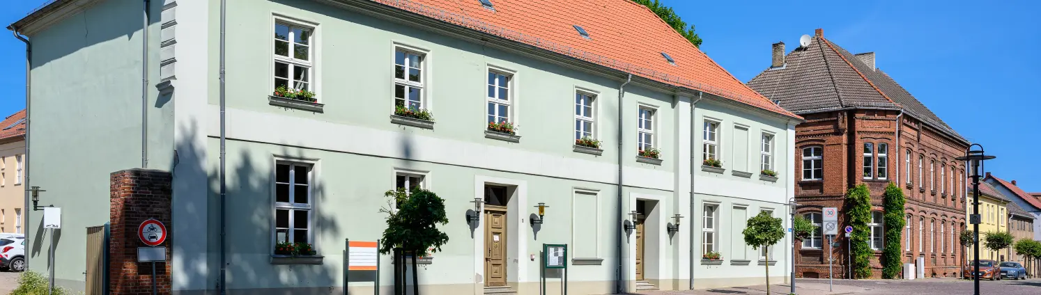 Das hellgraue Rathaus von Gransee in Brandenburg mit rotem Walmdach vor blauem Himmel.