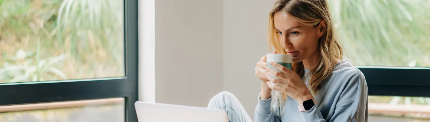 Frau sitzt in ihrer Wohnung und schaut auf ihren Laptopbildschirm mit einer Tasse Kaffee in der Hand