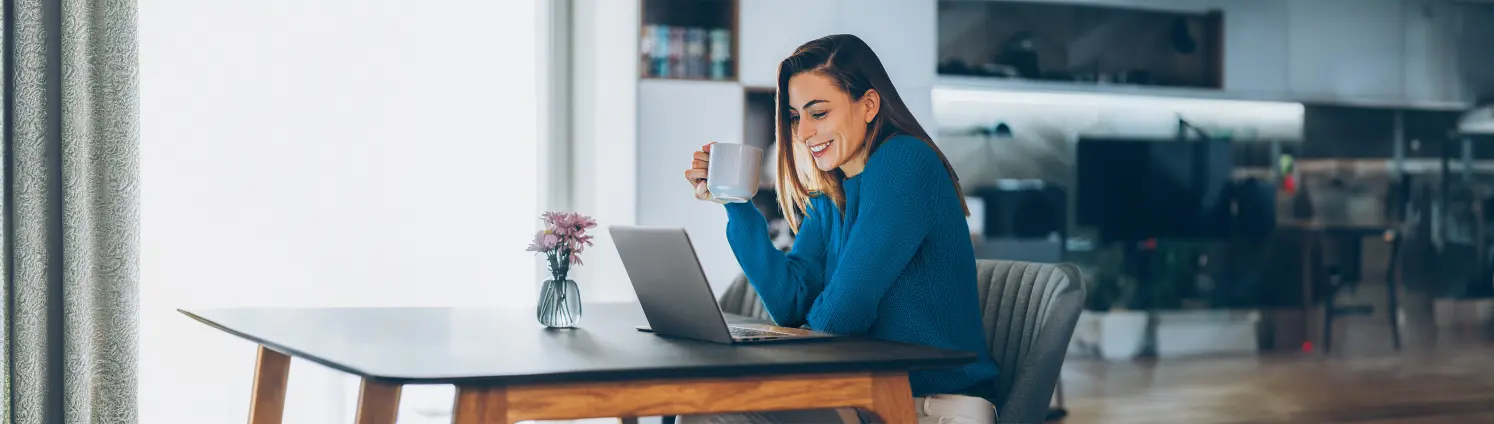 Eine junge Frau mit langem Haar sitzt am Tisch, blickt lächelnd auf ihren Laptop und hält eine Tasse in der rechten Hand.