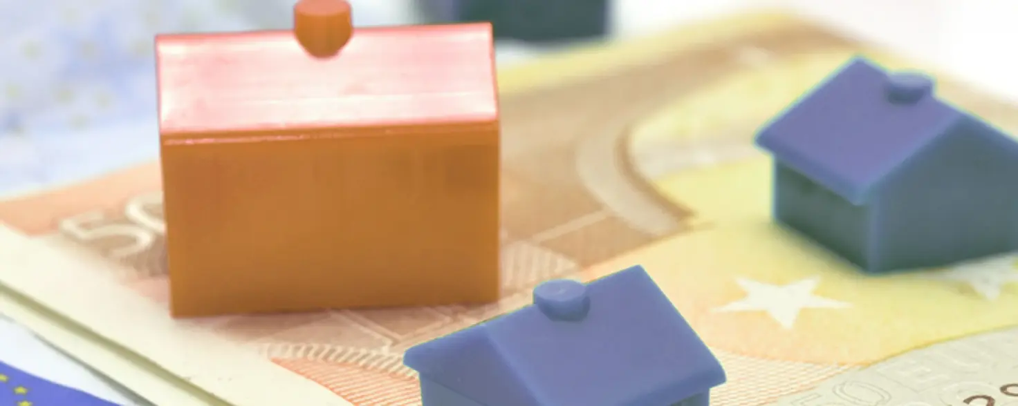Drei kleine blaue und rote Spielhäuser stehen auf einem hellbraunen Fünfzig-Euro-Schein. Darunter weitere blaue Geldscheine.