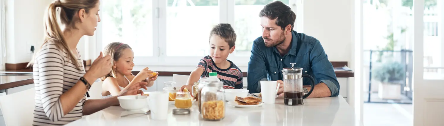 Mutter, Vater und zwei Kinder sitzen zum Frühstück an einem Tisch in einer hellen Küche. Es gibt Kaffee, Brote und Cornflakes.