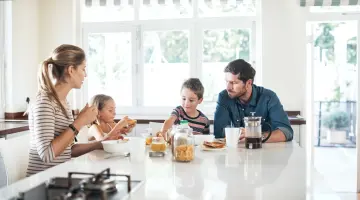 Mutter, Vater und zwei Kinder sitzen zum Frühstück an einem Tisch in einer hellen Küche. Es gibt Kaffee, Brote und Cornflakes.