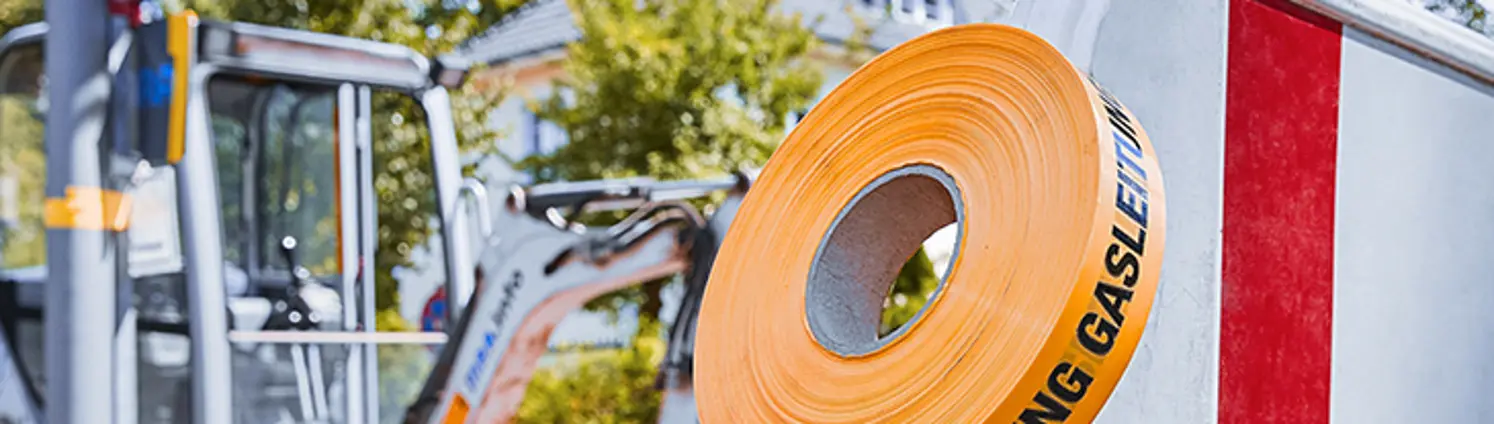 Eine Baustelle im Hintergrund. Im Vordergrunde eine rot-weiße Absperrung auf der ein Rolle Flatterband mit der Aufschrift 