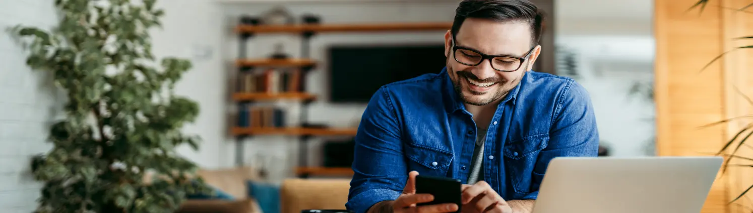 Ein dunkelhaariger Mann mit Brille und Jeanshemd sitzt am Schreibtisch. Sein Laptop ist geöffnet und er tippt lachend auf dem Smartphone.