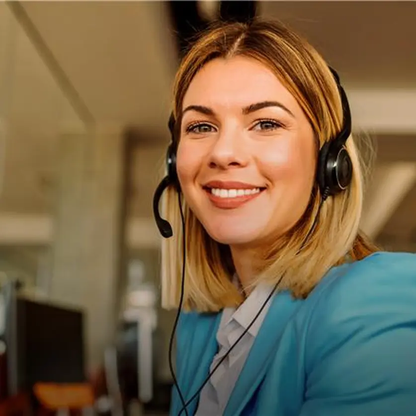 Blick in ein Kundenbüro: Eine junge Frau mit Headset sitzt vor dem PC und lächelt in die Kamera.