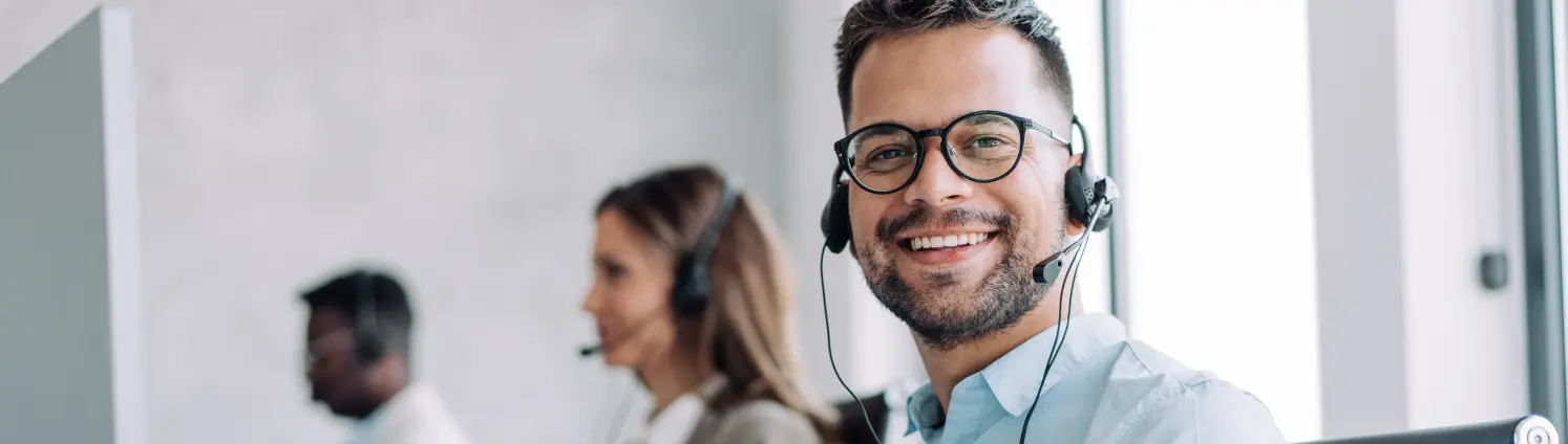 Blick in ein Kundenbüro: ein Mann mit Brille und Headset lächelt in die Kamera. Im Hintergrund Kolleginnen und Kollegen bei der Arbeit.