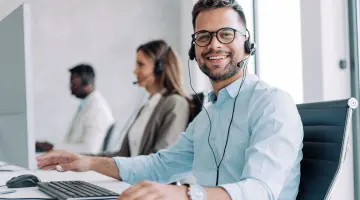 Blick in ein Kundenbüro: ein Mann mit Brille und Headset lächelt in die Kamera. Im Hintergrund Kolleginnen und Kollegen bei der Arbeit.
