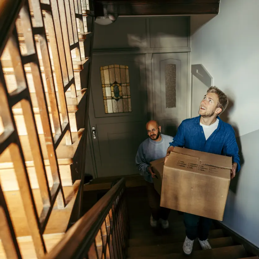 Zwei Männer tragen erwartungsfroh Umzugskisten die Treppen eines Altbaus hinauf in die neue Wohnung.