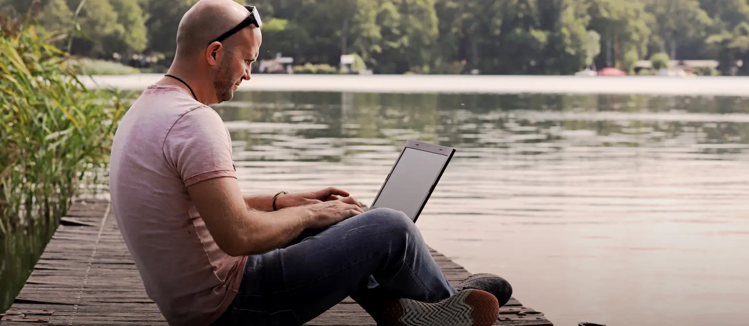 Ein Mann sitzt mit seinem Laptop auf einem Steg in einem See