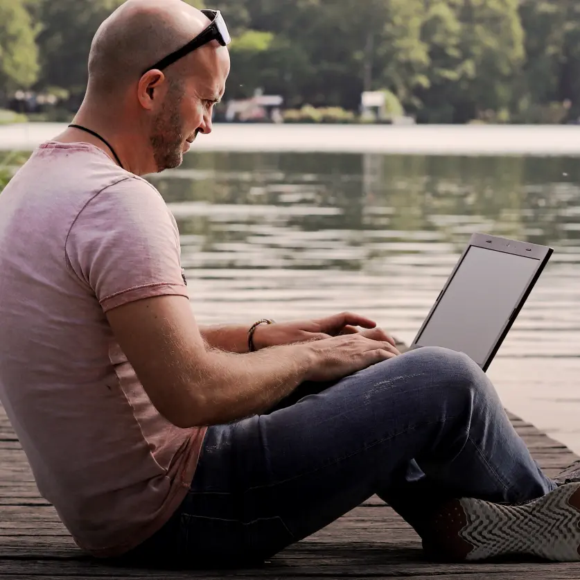 Ein Mann sitzt mit seinem Laptop auf einem Steg in einem See