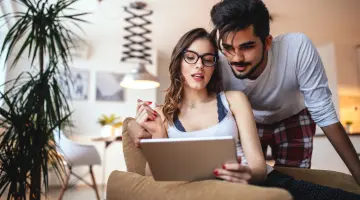 Eine junge Frau mit langem Haar sitzt am Tisch, blickt lächelnd auf ihren Laptop und hält eine Tasse in der rechten Hand.