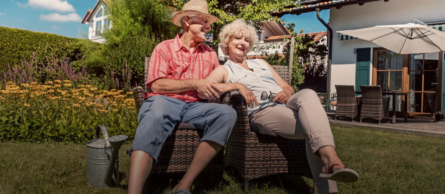 Sanierungsplan: Älteres fröhliches Paar sitzt im sonnigen Garten eines Einfamilienhauses mit Solaranlage auf dem Ziegeldach.