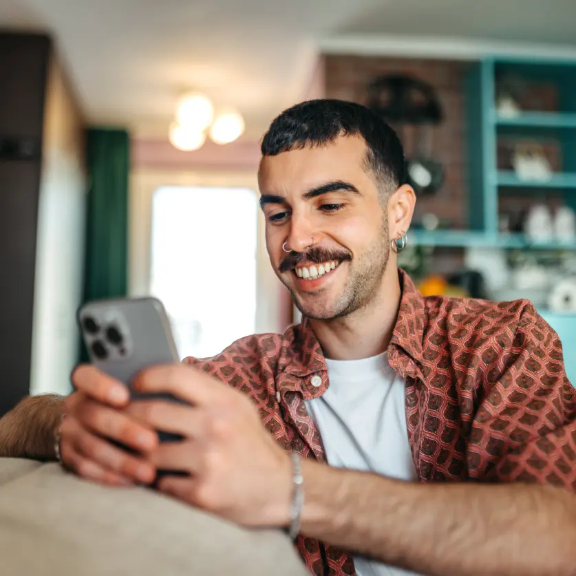 Junger Mann mit Nasenring und kariertem Hemd sitzt auf dem Sofa und tippt lächelnd auf seinem Smartphone.