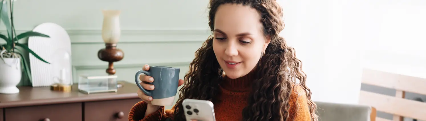 Eine junge Frau mit Tasse in der Hand sitzt im Sessel schaut auf ihr Smartphone.