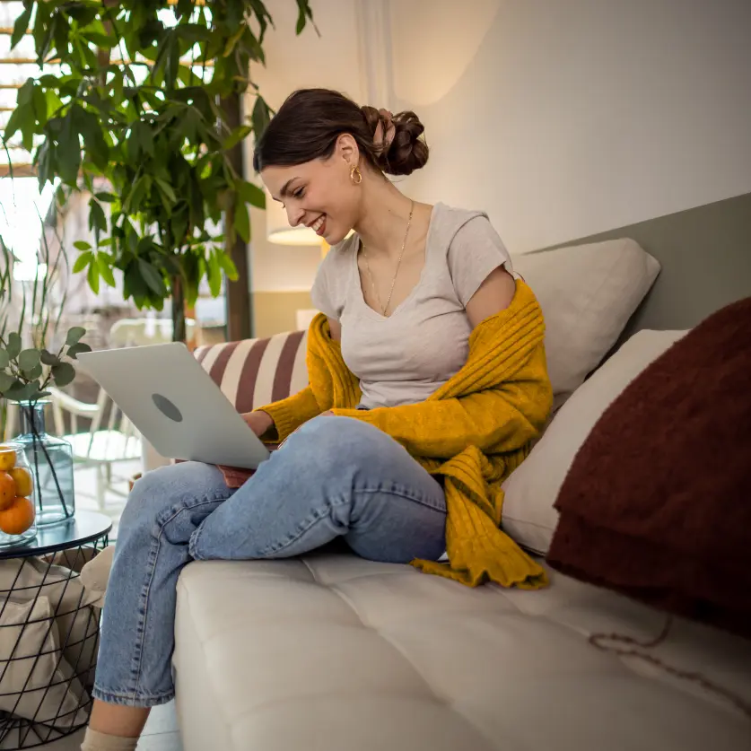 Lächelnde Junge Frau mit ockerfarbener Strickjacke sitzt mit hochgezogenem Knie auf dem Sofa und tippt am Laptop.