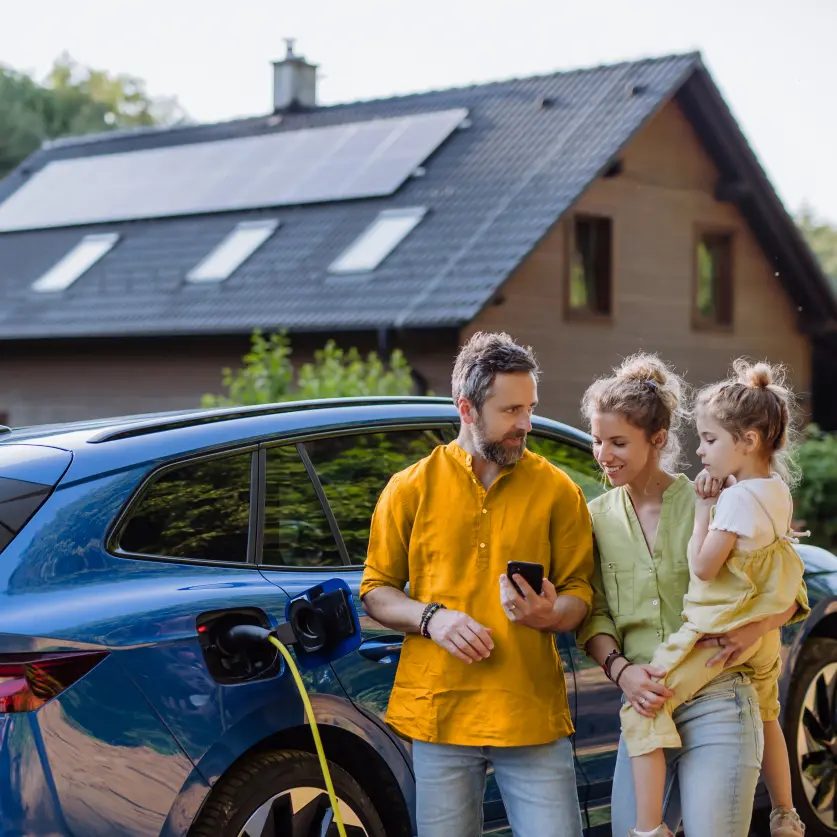 Familie mit Kind schaut auf ein Smartphone. Im Hintergrund ein Einfamilienhaus mit Solaranlage und ein blaues E-Auto das per Stecker geladen wird.