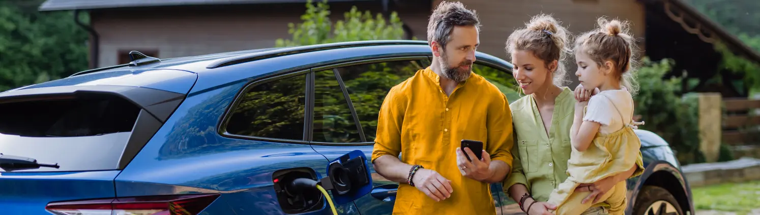Familie mit Kind schaut auf ein Smartphone. Im ein Einfamilienhaus mit Solaranlage und ein blaues E-Auto das per Stecker geladen wird.