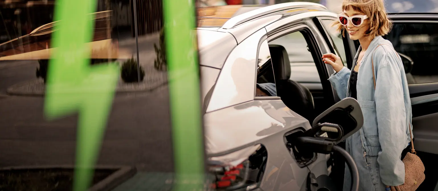 Junge Frau mit Sonnenbrille und Jeans lädt ihr Elektroauto an einer Ladesäule, die mit einem grünen Blitz bedruckt ist.
