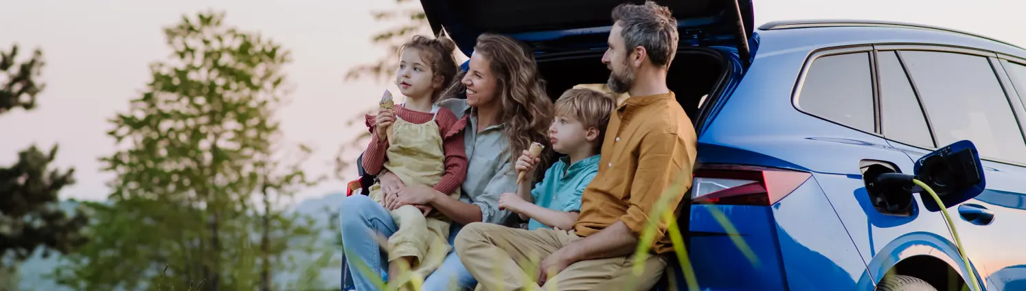 Fröhliche Familie, die im geöffneten Kofferraum ihres Elektroautos sitzt, Eis isst und auf das Aufladen des Akkus wartet.