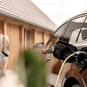 Eine schwarze Limousine lädt an einer Wallbox. Im Hintergrund betritt ein Mann mit Schultertasche ein Einfamilienhaus mit rotem Dach.