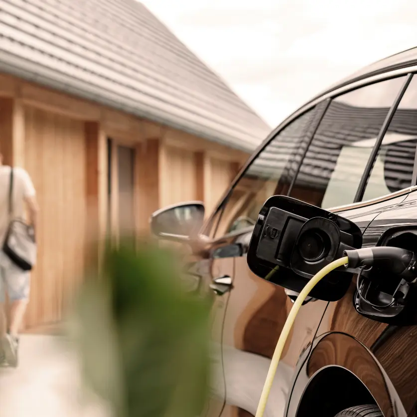 Eine schwarze Limousine lädt an einer Wallbox. Im Hintergrund betritt ein Mann mit Schultertasche ein Einfamilienhaus mit rotem Dach.