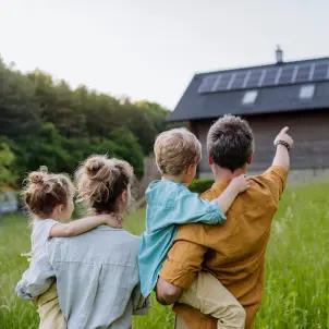 Ein Ehepaar mit zwei kleinen Kindern auf dem Arm steht auf einer Wiese. Der Vater zeigt auf ein Einfamilienhaus mit Solaranlage auf dem Dach.
