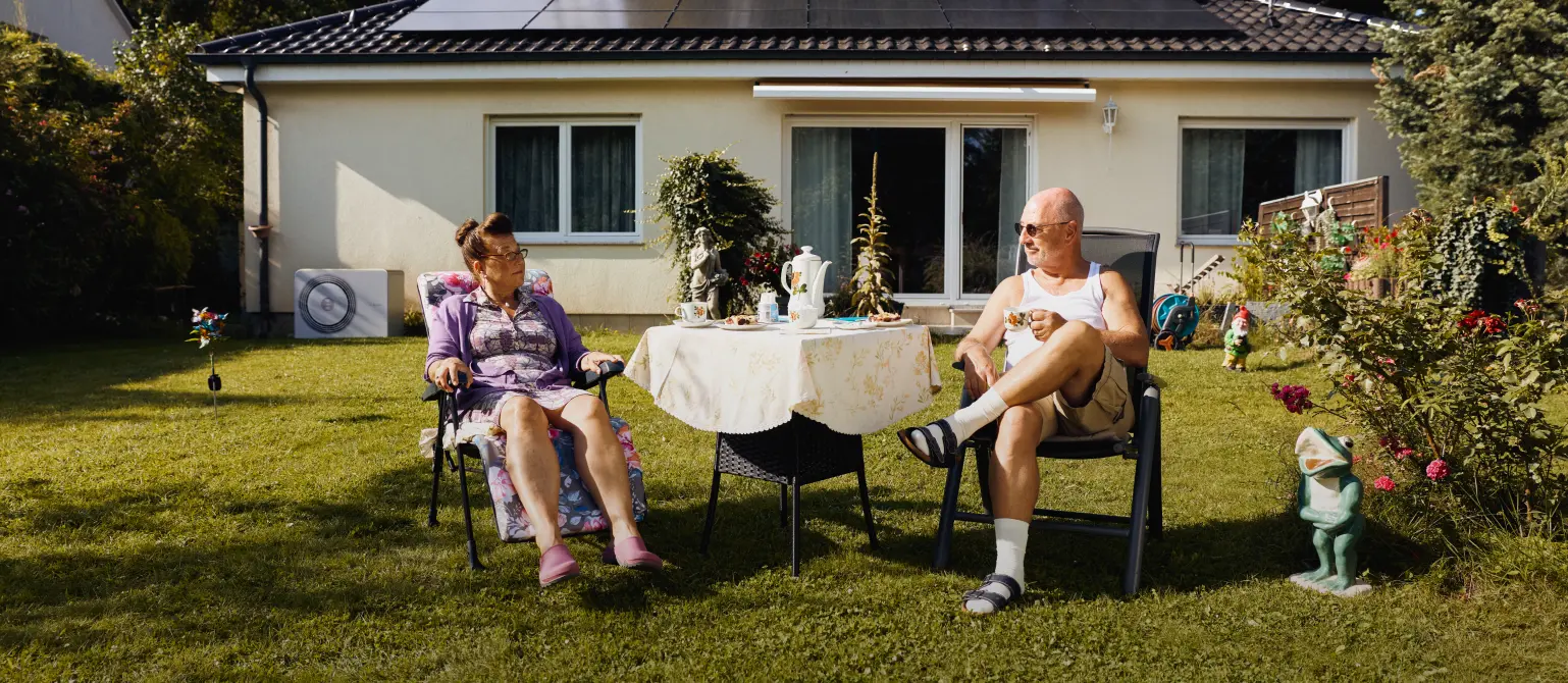 Ein älteres Ehepaar sitzt mit Sonnenbrillen am Gartentisch, umgeben von Gartenzwergen. Im Hintergrund ein Bungalow mit Solaranlage auf dem Dach.