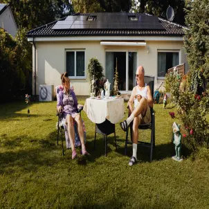 Ein älteres Ehepaar sitzt mit Sonnenbrillen am Gartentisch, umgeben von Gartenzwergen. Im Hintergrund ein Bungalow mit Solaranlage auf dem Dach.