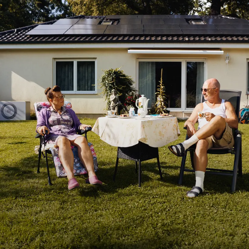 Ein älteres Ehepaar sitzt mit Sonnenbrillen am Gartentisch, umgeben von Gartenzwergen. Im Hintergrund ein Bungalow mit Solaranlage auf dem Dach.