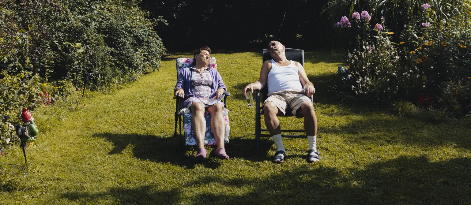 Ein älteres Ehepaar sitzt auf gepolsterten Gartenstühlen in der Sonne auf dem Rasen und macht ein Nickerchen.