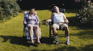 Ein älteres Ehepaar sitzt auf gepolsterten Gartenstühlen in der Sonne auf dem Rasen und macht ein Nickerchen.